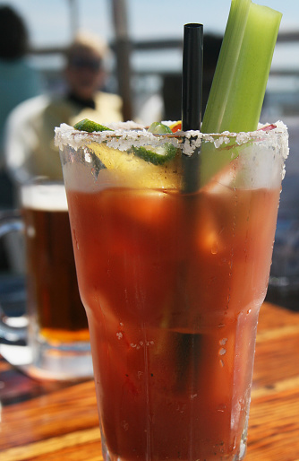 Bloody Mary drink in a frosted glass, complete with lime and celery, sits on patio table at California seaside cafe. Drink consists of vodka, tomato juice and spices. A straw is placed in the alcoholic drink. The rim is salted. 