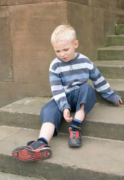 a five year old boy sitting looking down at his painful ankle or foot