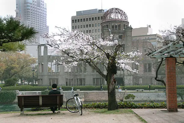 "A business man, his bicycle and cherry blossoms in the forground with the Hiroshima Atomic Bomb Dome (Gembaku Domu, formerly the Industrial Promotion Hall) in the background. The Aioi River runs between them. Overcast day.This was one of a very few buildings left standing within a two kilometer radius of the explosion. Amazing because the shock wave from the bomb created a pressure of 35 tons per square meter at the hypocenter."