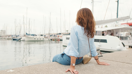 Attractive slim middle-aged woman sitting in the port looks into the distance at the yachts, back view