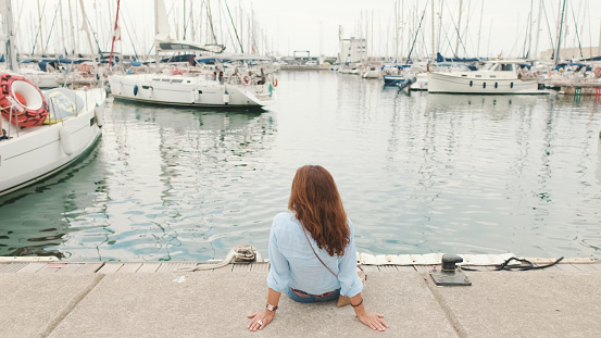 Attractive slim middle-aged woman sitting in the port looks into the distance at the yachts, back view