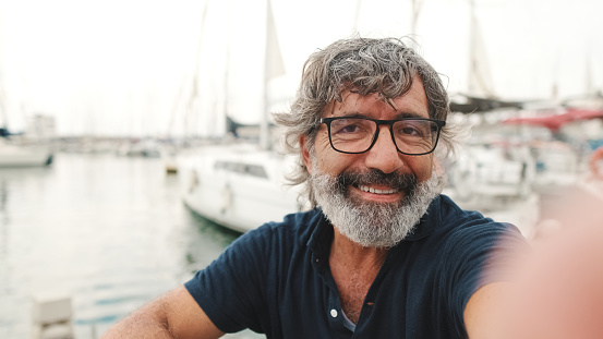 Close up, happy middle-aged man in glasses sits in the port on yacht background uses smartphone, makes selfie on mobile phone