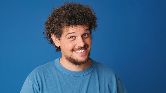 Close up, pensive guy with curly hair dressed in blue t-shirt looks at the camera having good idea on blue background in the studio