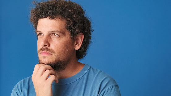 Close up, guy with curly hair dressed in blue t-shirt, thinks over his problem, remembering something isolated on blue background in the studio