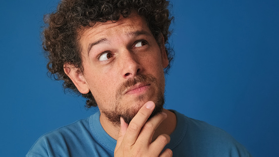 Close up, guy with curly hair dressed in blue t-shirt is thinking over difficult choice, imagining plan in his mind, having an idea, isolated on blue background in the studio