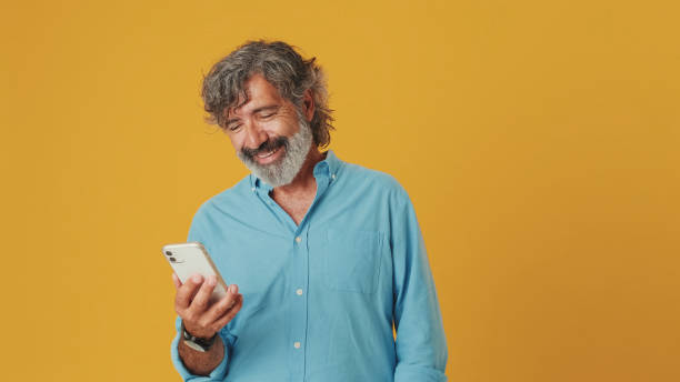 istock Happy elderly grey-haired bearded man wears blue shirt, uses mobile phone, having fun with mobile device, isolated on orange background in studio 1722657354