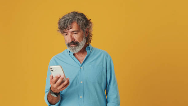istock Smiling elderly grey-haired bearded man wears a blue shirt, uses a mobile phone, texts, chats, isolated on an orange background in the studio 1722657356