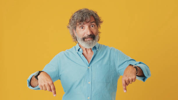 istock Elderly grey-haired bearded man wears blue shirt, pointing fingers down on workspace area copy space mock up, isolated on orange background in studio 1722659055
