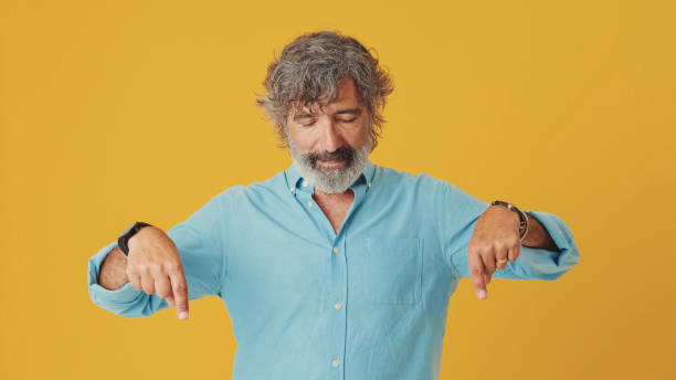istock Elderly grey-haired bearded man wears blue shirt, pointing fingers down on workspace area copy space mock up, isolated on orange background in studio 1722659058
