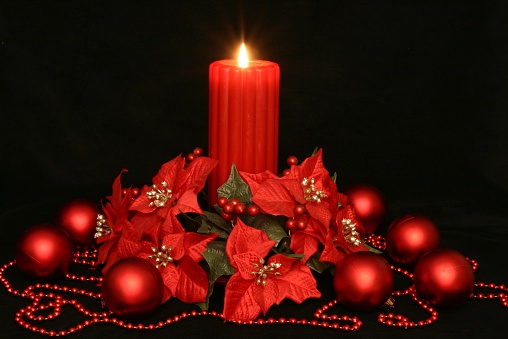 Vibrant red poinsettias & Christmas ornaments or baubles surround a burning red pillar candle on black background. Horizontal Christmas image.