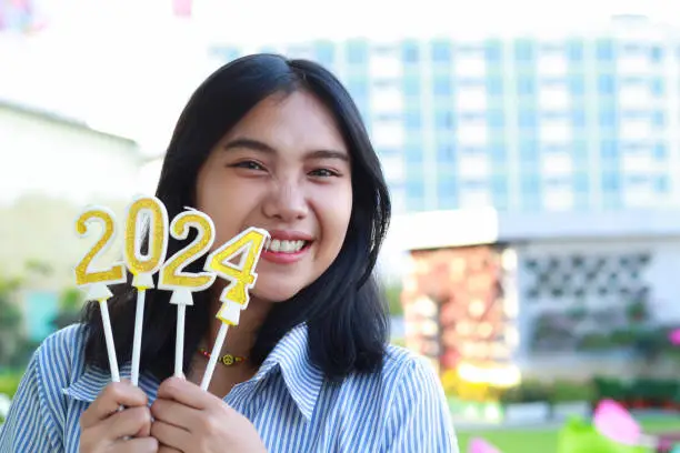 Photo of beautiful asian young woman looking to camera with excitement and holding golden candle number 2024, stylish female standing in apartment roof top with urban building background