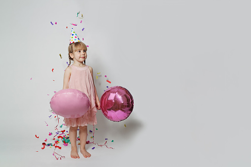 Happy little child girl in birthday hat having fun, holding balloon and standing in confetti rain on gray background