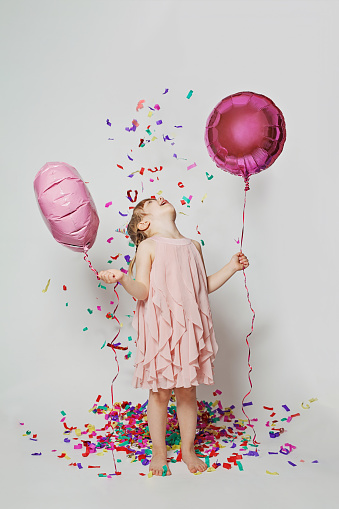 Happy child girl standing with balloon in colorful confetti rain and having fun on white color wall background with confetti. Birthday party concept