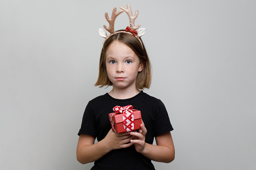 Small girl Christmas child in Xmas hairband standing with red present box on white background. Kid 7 years old