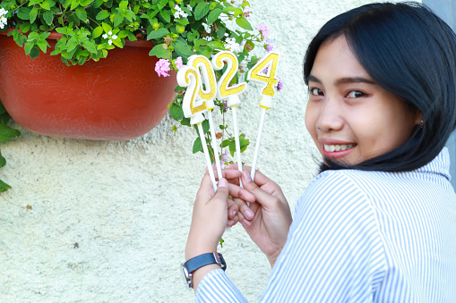 close up happy asian young woman smiling and looking to camera with holding 2024 number candle, elegance female standing in outdoor over vintage house with stone wall and green plant