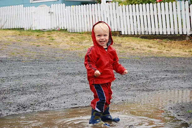 A young boy grins as he splashes in a rain puddle.  