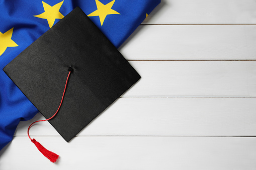 Black graduation cap and flag of European Union on white wooden table, flat lay. Space for text