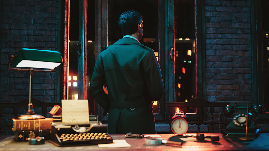 Detective wearing trench coat stands in his office at night looking out of a window.  Retro view or film noir look at the life as an detective. On his desk is a typewriter, a gun and an old phone.