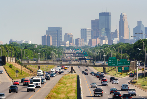 The busy multiple lane highway freeway that leads into the city of Minneapolis, Minnesota. The summer daytime urban scene shows heavy traffic of cars and trucks with the distant downtown city skyline in the background. Hazy blue sky area provides copy space.