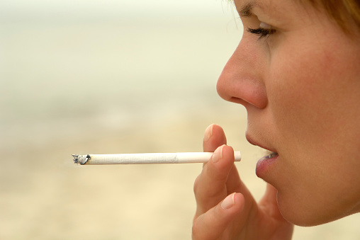Close-up of woman's face smoking slim cigarette, sand beach in the background.