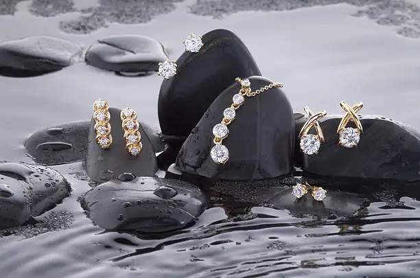 An assortment of gold diamond earrings with an eternity necklace laying upon a bed of black rocks and rippled water.