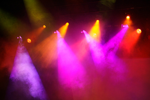 Approximately ten orange and pink bright stage spot lights shinning from above the empty stage. The lights are hanging and the frame supporting them is slightly visible. The stage lights are pointed downward. There is fog in the air creating richer tones from the lights. 