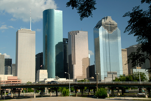 Horizontal image of modern skyscrapers in Houston, Texas, skyline against the blue sky with clouds.  Silhouetted tree branches with leaves, partially frame the shot in the upper right corner.