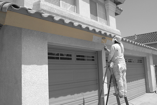 Worker rolls paint onto the trim of an average American house. The entire house, which has been prepped for paint, is obviously black and white while the freshly rolled paint has been left its original brown color which draws attention to the job at hand. Canon 30D, 28-55mm lens.