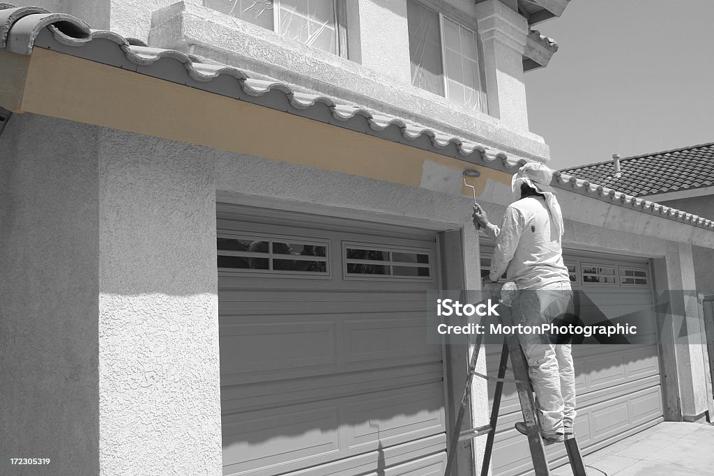 Painter on a stool painting the building from white to beige Worker rolls paint onto the trim of an average American house. The entire house, which has been prepped for paint, is obviously black and white while the freshly rolled paint has been left its original brown color which draws attention to the job at hand. Canon 30D, 28-55mm lens. Repairing Stock Photo