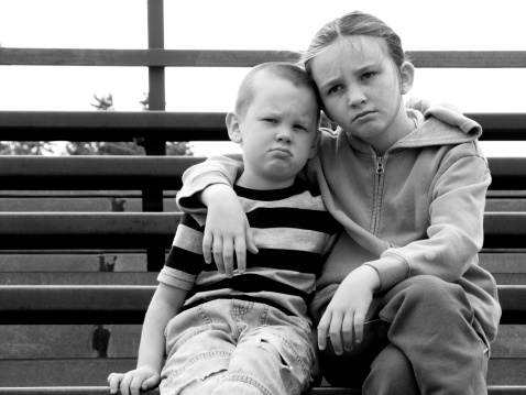 Siblings sitting on wooden bench look with upset and disappointed expressions. Brothers sit on different sides of bench in silence after argument
