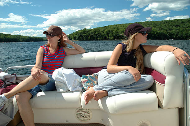 Mother And Daughter Enjoying Pontoon Boat On Lake "Horizontal image of a mother and daughter enjoying a ride on the lake while relaxing on the soft cushions of a pontoon boats' seating.  A beautiful blue sky with clouds , the lake , and tree covered hills are in the background." pontoon boat stock pictures, royalty-free photos & images