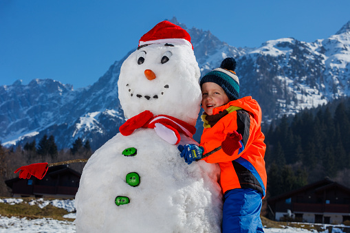 Handsome little boy in winter sport outfit play with snowman in Santa hat outside