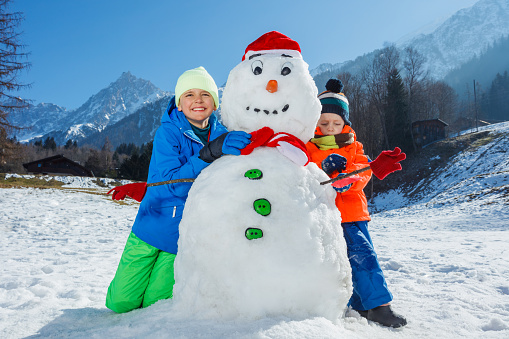 Portrait of two happy boys brothers in winter sport outfit play with snowman in Santa hat and scarf outside