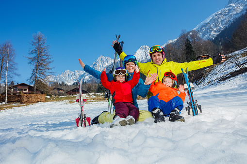 Family, father son and little kids sit in the snow in sport outfit with skis helmets masks laugh hug smiling over mountain