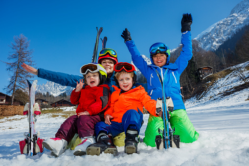 Fun on first ski alpine vacation family of mom and three kids sit hugging in snow wear helmets sport outfit masks lift hands up