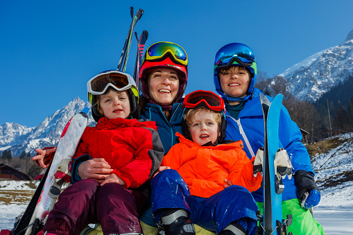 Close-up family portrait of four sit in snow, mother,boy and two little kids dressed up in sport outfit helmets masks, laughing hugging smiling enjoying the beautiful mountain view