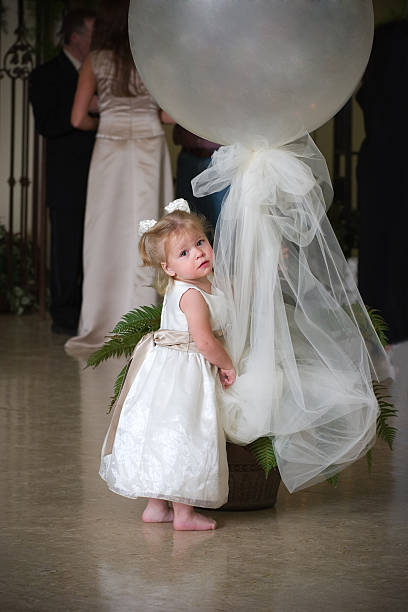 Cute Barefoot Child Wearing White Dress Holding Balloon Decoration Cute little girl wearing a white dress with ribbons in her hair, holding onto ribbon of a large decorative party balloon. flower girl stock pictures, royalty-free photos & images