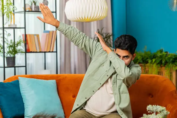 Photo of Happy man having fun, dancing, dabbing raising hands, making dub dance gesture at home room on sofa