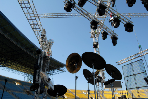 A collection of drummer's cymbals stands on the stage of a stadium concert setup. Shot from below against a clear blue sky with various pieces of lighting equipment and truss in the background.
