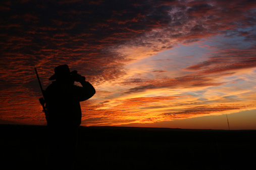 A male hunter in a cowboy hat scans the terrain at sunset. 