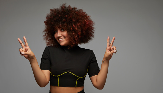 A young woman with curly hair, afro and mulatto, making a peace sign with both hands.