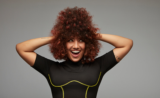 A smiling, mixed race young woman with her hands in her fluffy curly hair.
