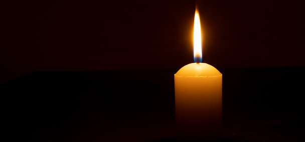 Single burning candle flame or light is glowing on a big white candle on black or dark background on table in church for Christmas, funeral or memorial service with copy space.