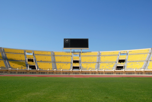 A scoreboard with a clock sits atop yellow seats in an empty stadium. Shot against a clear blue sky with nice green grass in the foreground.
