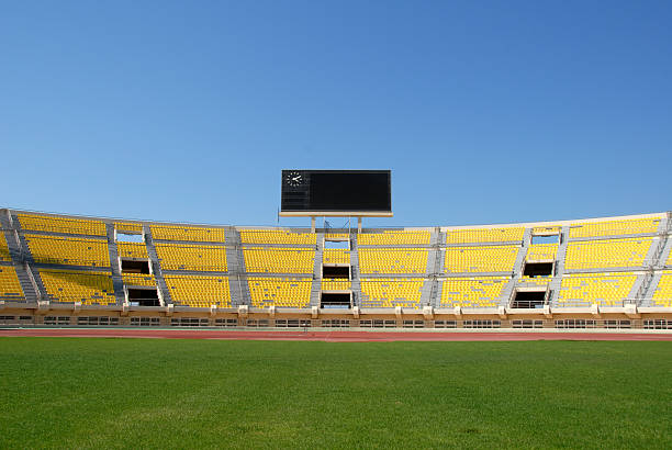 Scoreboard in Empty Stadium A scoreboard with a clock sits atop yellow seats in an empty stadium. Shot against a clear blue sky with nice green grass in the foreground. scoreboard stadium sport seat stock pictures, royalty-free photos & images
