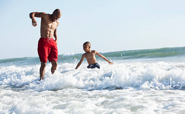 Father and son playing in surf Black father (30s) and son (6 years) at the beach, playing in the surf. waist deep in water stock pictures, royalty-free photos & images