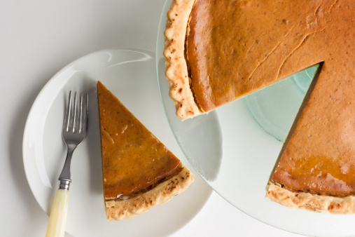 A pumpkin pie on a glass serving platter stand. A wedge is cut out of the round pastry, and the removed slice piece is on a plate with a fork next to it, ready to serve for Thanksgiving holiday dessert. On a white background.
