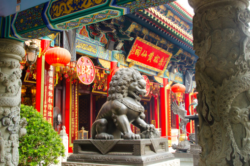 November 13, 2023. Wong Tai Sin Temple - Wong Tai Sin area, Hong Kong. Black color statue of lion dog in front of Wong Tai Sin temple's main building decorated with red color ribbon during the day
