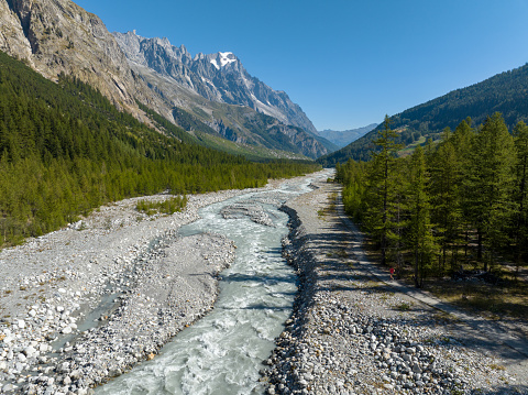 An aerial shot of a fit mid adult woman running along a pathway between a lush green pine forest and a glacial river. Woman running in nature to keep active and healthy.