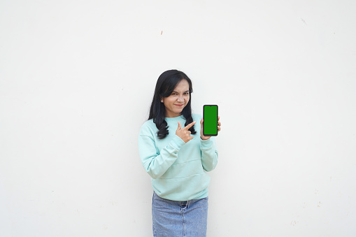 Portrait Indonesian beautiful woman wearing light blue sweater with happy and smile face show green screen mobile phone isolated on white background